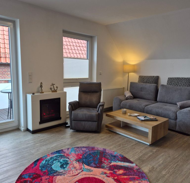 „Wohnbereich der Ferienwohnung Tillmanns Ankerplatz in Norden mit Kamin, Relaxsessel, Sofa und neuem Teppich.“